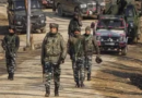 Group of soldiers patrolling a rural road with military vehicles in the background.