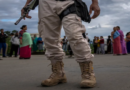 A person in military attire holding a weapon stands in front of a group of people on a cloudy day.