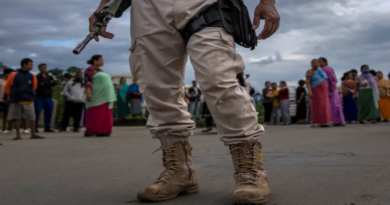 A person in military attire holding a weapon stands in front of a group of people on a cloudy day.